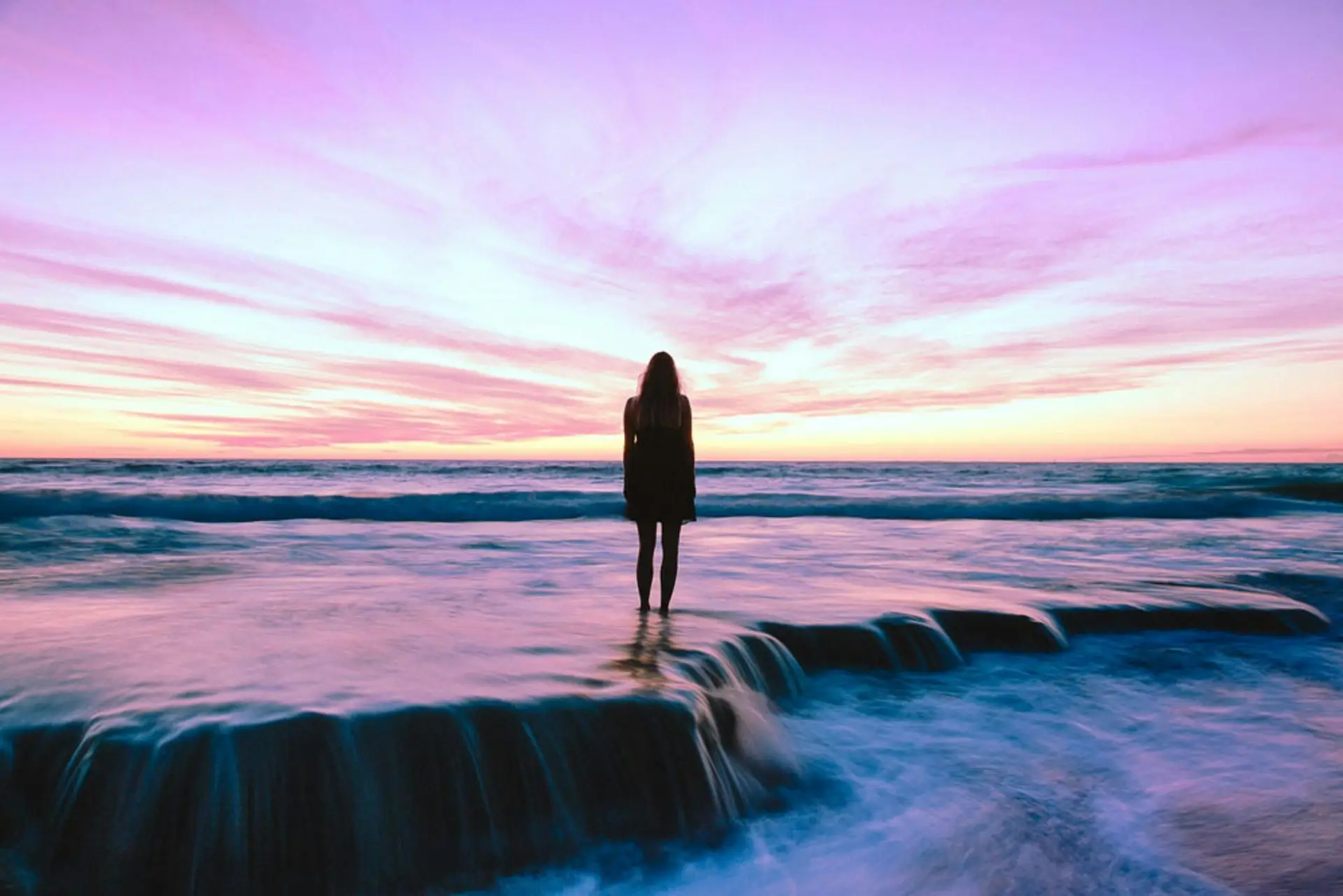 A woman stands alone by the ocean at sunset, embracing nature's beauty.